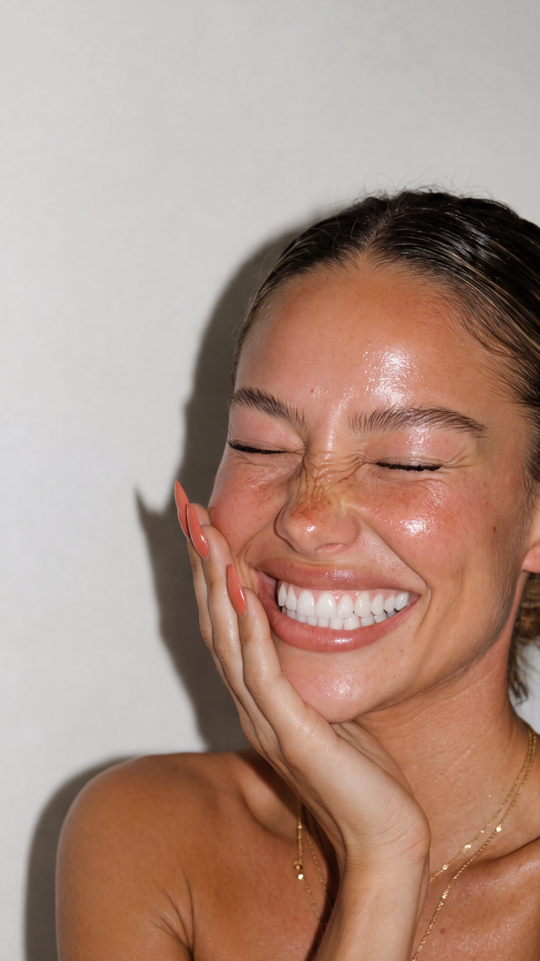 Close-up portrait of a smiling woman with glowing skin, closed eyes, and her hand resting against her cheek.